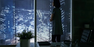 美丽的女商人从她的私人办公室的窗户看向大城市，然后坐在她的办公桌。她的工作空间用暗色调看起来很现代。