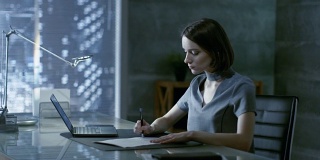 成功的年轻女性坐在私人办公室的办公桌前，用笔记本电脑工作，签署重要文件。她的办公室是极简主义风格，相当黑暗，有窗户可以看到大城市的景色。