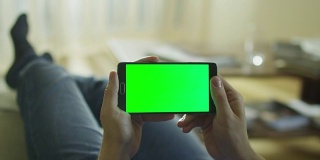 一名男子躺在家里的沙发上，手里拿着安卓手机，绿屏是横屏模式。
