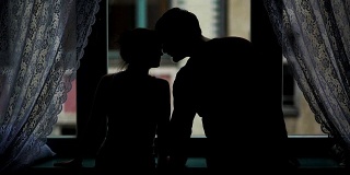 窗户附近一对夫妇的剪影肖像。女人牵着男人的手，充满爱意地亲吻和拥抱。《黑暗中的年轻家庭》的后视图
