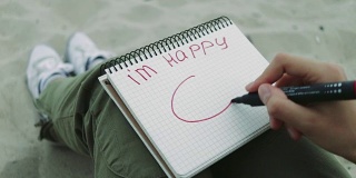 晚上，女孩在沙滩上用红色毡尖笔在笔记本上写着“我很快乐”，并画了一个笑脸