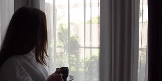 年轻女子长头发在白色浴袍喝咖啡站在窗户附近在家。