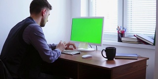 一个年轻人正在电脑上工作，窗户旁边的桌子上有一个绿色屏幕的模型。白人工人在键盘上打字，看着绿色的屏幕