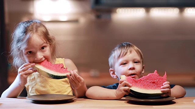 哥哥和妹妹坐在厨房的桌子旁。男孩和女孩吃着多汁的西瓜，看着镜头