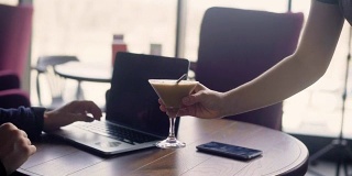 一个服务员用托盘端着一杯鸡尾酒，给一个在餐厅里通过笔记本电脑上网工作的忙碌男士