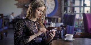 一位女士用信用卡和智能手机在网上购物坐在餐馆里，后面有人。一个穿着黑色裙子的女人用现代手机进行无现金支付，并在网上购物