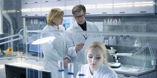走过现代实验室并讨论未来可能性的女性和男性首席科学家。年轻的女助理在她的办公桌前工作。