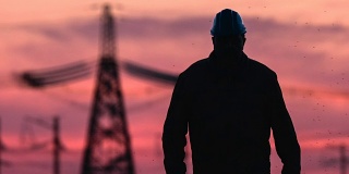 中景监工剪影在安全帽沿着电线在红色日落期间能源变电站检查。