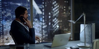 商人坐在办公桌前，在办公室里讲电话，可以看到大城市的风景。