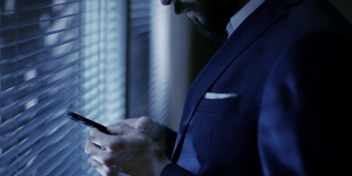 深夜，一个商人站在他私人办公室的窗户前使用智能手机。