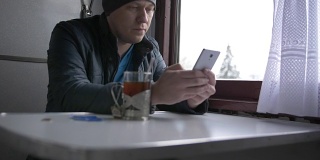 一位戴着灰色帽子、穿着蓝色夹克的年轻人手持白色智能手机与人交流，微笑着将智能手机放在桌子上，然后端上一杯茶