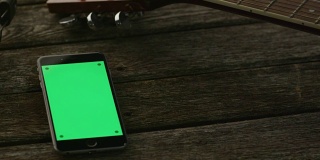 肖像模式下的绿色屏幕手机放在吉他和耳机旁边的木桌上。因果的生活方式