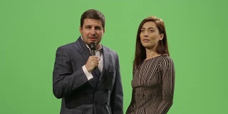 媒体记者正拿着麦克风对一名身着连衣裙的女子进行采访，背景是绿色屏幕模型。