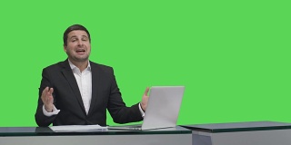 媒体广播员正坐在一张桌子旁，对着背景中的绿色屏幕模型说话。