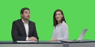 男女广播员坐在一张桌子旁，对着背景中的绿色模拟屏幕说话。