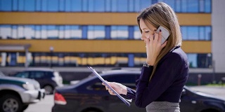 一位忙碌的商务女士拿着重要的合同，一边讲着手机，一边走过停着很多车的停车场
