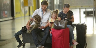 一家四口正带着大包大包等在火车站或机场。父母和孩子一起使用移动设备。旅游的概念