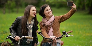 两名骑自行车的女性正在用智能手机拍照。年轻漂亮的黑发美女骑着自行车用手机给自己拍照