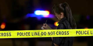 女亚裔美国女警察记录在犯罪现场与闪烁的警车警报器在背景