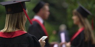 金发女孩穿着学位服，戴着毕业帽在智能手机上上网