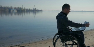 一个身体残疾的人坐在海边的轮椅上，手里拿着笔记本电脑