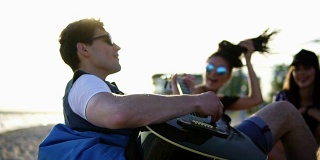 一个年轻人和一群朋友坐在沙滩上的安乐椅上弹吉他，在一个夏日傍晚的夕阳下唱歌。Slowmotion拍摄