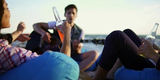 一群朋友坐在沙滩上的安乐椅上，喝着鸡尾酒和啤酒，欢呼雀跃，听着朋友在一个夏日傍晚的夕阳下弹吉他。Slowmotion拍摄