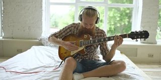 有魅力的大胡子男子坐在床上学习弹吉他，在家里的现代卧室里用平板电脑