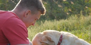 一个年轻人在户外抚摸、拥抱、亲吻他的拉布拉多犬。和金毛猎犬玩耍。狗舔男人的脸。与家畜的爱情和友谊。景观背景。Slowmotion