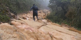年轻小伙子穿着雨衣，在岩石小道上蹦蹦跳跳。徒步旅行者背着背包在潮湿的森林里的石头地上奔跑。男游客走在丛林小径上。慢镜头后视图