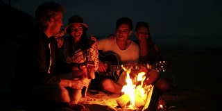 年轻人晚上在海滩上用篝火野餐。欢快的朋友们唱着歌，弹着吉他。Slowmotion拍摄