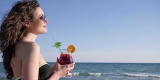 性感的女人与卷发饮料多彩的鸡尾酒，微笑的女孩在海滩上的肖像，闪亮的海背景