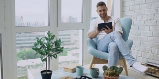 在阁楼现代公寓里，有魅力的男人坐在阳台的椅子上使用数码平板电脑
