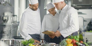 在现代厨房团队中，厨师们用平板电脑制作食谱，他们微笑着讨论。厨房里有很多食材，蔬菜，肉，汤。