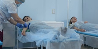 两名女病人躺在医院病房的床上打点滴，由两名医生检查