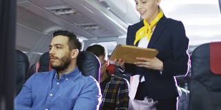 空姐向西班牙男性乘客展示带有菜单的平板电脑。他们在机上。商务舱的一个商业航空内部可见。