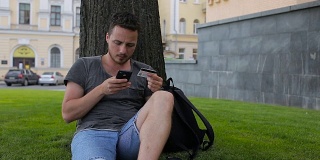 男子用信用卡在网上购物时用智能手机在公园的一棵树下坐着