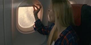 在飞机上，美丽的金发女孩打开窗帘向窗外望去。阳光照耀着她，她微笑着。
