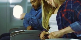 在一架飞机上美丽的金发女性和英俊的西班牙裔男性系好安全带，准备起飞/降落。阳光透过飞机的窗户。