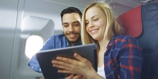 在商用飞机的一个板上，漂亮的年轻金发美女和英俊的西班牙男性使用平板电脑和微笑。阳光透过飞机窗户。