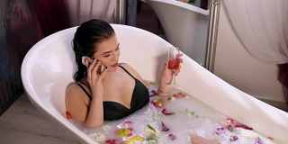 年轻漂亮的女人在打电话，在牛奶浴中放松，从杯子里喝起泡酒。在4 k拍摄