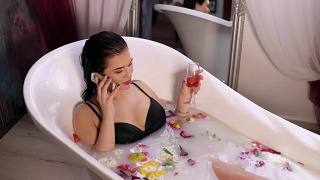 年轻漂亮的女人在打电话，在牛奶浴中放松，从杯子里喝起泡酒。在4 k拍摄视频素材模板下载