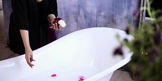 年轻迷人的女子在黑色浴衣扔花到浴缸装满牛奶，准备洗个热水澡。Slowmotion拍摄