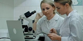 穿着实验服的研究人员在明亮的实验室里用显微镜检查样品，使用数码平板电脑，在笔记本电脑上做笔记