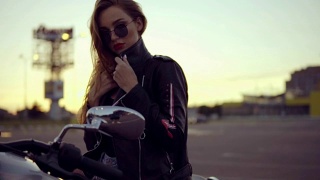 一个女自行车手的肖像，穿着皮夹克和短裤坐在她的自行车上，握着把手。年轻性感的卷发女人在直升机上戴着太阳镜的特写视频素材模板下载
