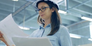 戴安全帽的女工程师站在大工厂的中央，绘制蓝图。