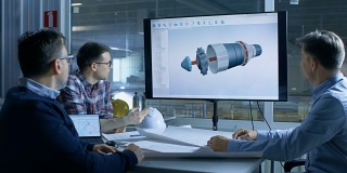 工业工程师小组讨论三维模型的涡轮/发动机设计显示在演示显示。在背景工厂被看到。
