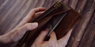 俯视图，一个女人在数，从她的棕色皮夹里拿出美元。女性拿着钱包，背景是木制的。慢动作