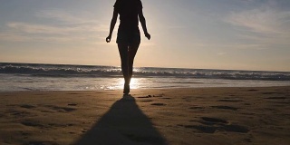一位年轻女子在日落时分跑到海边，举起手。美丽的年轻女孩在沙滩上去海边享受假期的自由。暑假放松一下。后方后视图