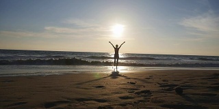 日落时分，一位年轻女子在海边的海滩上散步，她举起手。美丽的年轻女孩站在海边度假，享受自由。暑假放松一下。后视图慢动作